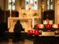 Candles glow in a church, with a sign reading "Prayer Intentions." People kneel in prayer, their mass intentions illuminated by the soft light.