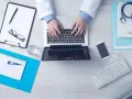Lekarz pisze na laptopie, obok stetoskop, recepta, telefon i termometr.