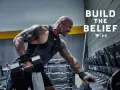Dwayne "The Rock" Johnson w koszulce Under Armour wykonuje ćwiczenia z hantlami. W tle stojaki z ciężarami.