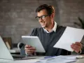 Uśmiechnięty mężczyzna w okularach analizuje dokumenty i tablet. Zastanawia się, jak długo jeszcze potrwa jego mały zus plus.