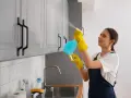 Kobieta w żółtych rękawiczkach czyści meble z okleiny, używając sprayu i ściereczki.