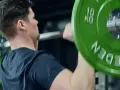 Man utför axelpress med skivstång. Gröna viktskivor med texten "SWEDEN" och "10 KG" syns tydligt.