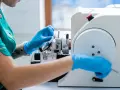 Ręce w niebieskich rękawiczkach pracują przy mikrotomie, przygotowując próbkę tkanki do badania. Może to być kluczowe dla diagnozy, czy wykryty guz jest złośliwy.