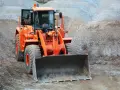 Pomarańczowa ładowarka kołowa z otwartymi drzwiami kabiny stoi na placu budowy, na tle hałd żwiru.