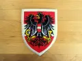 Naszywka z godłem Austrii: czarny orzeł z tarczą w biało-czerwone pasy, trzymający sierp i młot.