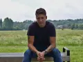 Bartłomiej Bis siedzi na ławce w parku, w tle zielone drzewa i niebo.