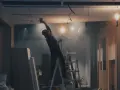 Budowlaniec na drabinie montuje sufit podwieszany, w tle widać materiały budowlane i wiszące kable.