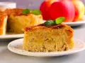 Una fetta di torta di mele vegana, soffice e ricca di mele, guarnita con menta. Sullo sfondo, una torta intera e mele rosse.