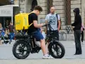 Młody kurier Glovo na rowerze elektrycznym sprawdza telefon. Obok stoi mężczyzna, a w tle widać ludzi i budynki.