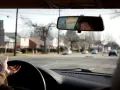 Mano sul volante, pronta per una manovra di svolta. Riflesso del volto nel retrovisore, case e alberi spogli fuori dal finestrino.