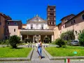 Kobieta w niebieskiej sukience pozuje przed kościołem Santa Cecilia w Trastevere. W tle wieża i fontanna.