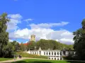 Ein sonniger Tag im **Rudolph Wilde Park**. Ein langer, weißer Pavillon spiegelt sich im Wasser, während ein Uhrenturm über Bäumen aufragt.