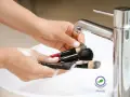 Mani che lavano pennelli da trucco insaponati sotto un rubinetto cromato. Impara come pulire i pennelli da trucco per una pelle sana.