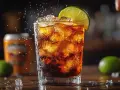 Szklanka z Cuba Libre, lodem i plasterkiem limonki, w tle butelka i puszka.
