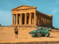 A lone traveler explores the Valley of the Temples in Sicily, standing before an ancient Greek temple and a fallen bronze statue.