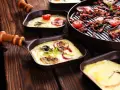 Raclette co to? Roztopiony ser z dodatkami w małych patelenkach i grillowane mięso z pomidorkami na drewnianym stole.