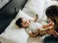 Uśmiechnięte dziecko bawi się zabawką na łóżku. Mama patrzy na malucha. Co to za mastka?