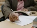 Osoba pisze ręcznie notatki w zeszycie, przygotowując list motywacyjny erasmus. W tle widać otwartą książkę.