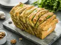 Pyszny chlebek rwany z pesto, udekorowany listkami bazylii, prezentuje się apetycznie na kamiennej desce. Obok czosnek i orzechy włoskie.