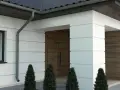 Nowoczesny dom z białym boniowaniem, drewnianymi drzwiami i ciemnym dachem.