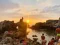 Tramonto dorato sulla piscina di Venere a Milazzo. Persone sedute sulle rocce e in acqua, godendosi la vista.