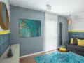 Sala de estar con sofá gris, alfombra turquesa y arte abstracto en tonos fríos. Un espejo redondo de madera y detalles en amarillo complementan el espacio.
