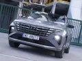 Szary Hyundai Tucson hybryda z charakterystycznym grillem i światłami LED, zaparkowany przed zielonym ogrodzeniem.