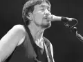 Chris Rea nie żyje. Muzyk śpiewa do mikrofonu na czarnym tle, w czarnej koszulce bez rękawów.