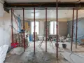 Chantier d'une maison phenix 1970 : poutres métalliques, étais, tuyaux et matériaux de construction dans une pièce en rénovation.