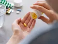 Dłoń wysypująca żółte tabletki z pojemnika na drugą dłoń, obok szklanka wody i opakowania leków.