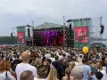 Tłum ludzi na koncercie pod gołym niebem. Na scenie gra zespół, a na ekranach wyświetlany jest green festival line up.