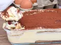 Un cucchiaio preleva una porzione cremosa di tiramisù, con 500 g di mascarpone, cosparso di cacao. Sullo sfondo, uova fresche.
