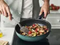 Ręce mieszają warzywa na patelni: pieczarki, pomidorki, bakłażan.