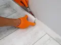 Ręce w pomarańczowych rękawiczkach przycinają listwę przypodłogową, przygotowując ją do wymiany panela podłogowego.