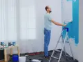 Mężczyzna maluje ścianę na niebiesko wałkiem, stojąc na drabinie.