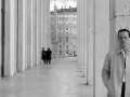 Un uomo in trench osserva l'obiettivo, mentre sullo sfondo due figure in abito scuro passeggiano sotto un colonnato. Potrebbe essere una scena da "Il talento di Mr. Ripley dove è stato girato".