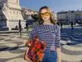 Dziewczyna w okularach i bluzce w paski, z kolorową torbą, nawiązuje do mody lat 60. na tle miejskiego placu.