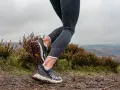 Gambe di una donna che fa trail run su un sentiero fangoso, con erica e colline nebbiose sullo sfondo.