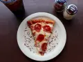 Kawałek pizzy pepperoni na talerzu, posypany płatkami chili i parmezanem. Idealny na szybki lunch, choć warto pamiętać o jego kcal.
