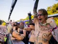 Feiernde Menschenmenge bei einem Festival, die zu **techno music** tanzt. Ein Mann mit Tattoos und Sonnenbrille jubelt.