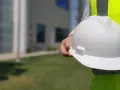 Biały kask w dłoni pracownika na budowie, obok jaskrawożółtej kamizelki. Różne kolory kasków na budowie zapewniają bezpieczeństwo.