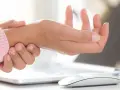 En person håller om sitt handled, kanske för att lindra smärta med ett elastiskt handledsstöd, vid en dator.