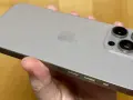 A hand holds a silver iPhone 15 Pro Max, showing its triple camera system and Apple logo.