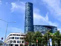 Wysoki, szklany budynek Sky Tower we Wrocławiu, z pewnością ma wiele pięter. Widać też niższy budynek i drzewa.