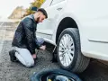 Mężczyzna klęczy przy białym samochodzie, używając podnośnika, by podnieść auto bez kanału. Obok leży zapasowa opona.