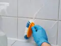 Rękawiczka w niebieskiej rękawicy szoruje szczotką fugi między płytkami, które często stają się problematyczne.