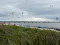Kitesurfare förbereder sig vid Habo Ljung Camping. Många kites svävar i luften över en strand med en lång pir i bakgrunden.