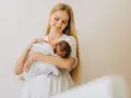 Uśmiechnięta młoda mama tuli noworodka, karmiąc go piersią. Delikatne spojrzenie i biała sukienka podkreślają jej macierzyństwo.