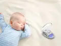 Śpiące dziecko w niebieskim pajacyku obok otwartego etui z monitorem oddechu dla niemowląt.