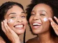 Deux femmes souriantes appliquent une crème sur leur visage, un geste pour une exfoliation visage réussie.